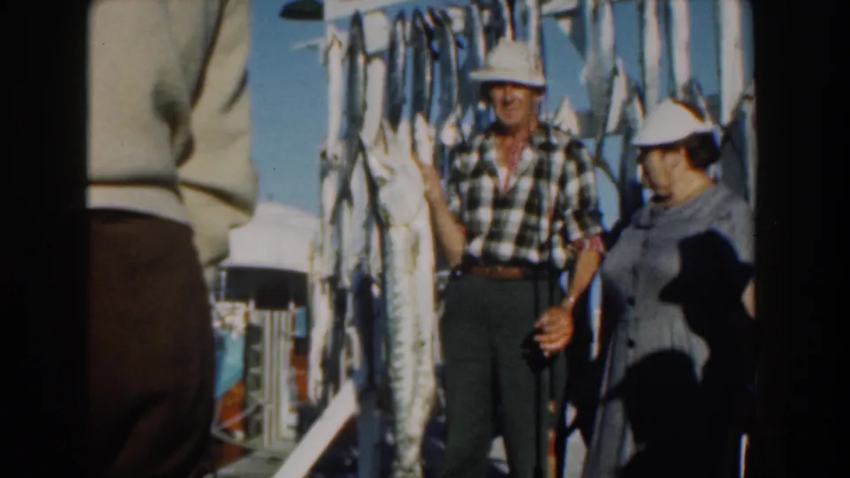 In Nassau, Bahamas in 1961, a couple poses on a boat beside large hanging fish, with blue sky and boat fittings behind them.