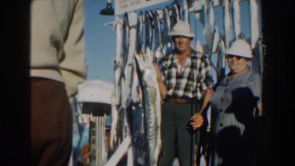 The couple poses in bright Nassau sunlight beside several large fish hanging from the boat structure, with soft 8mm color and framing.