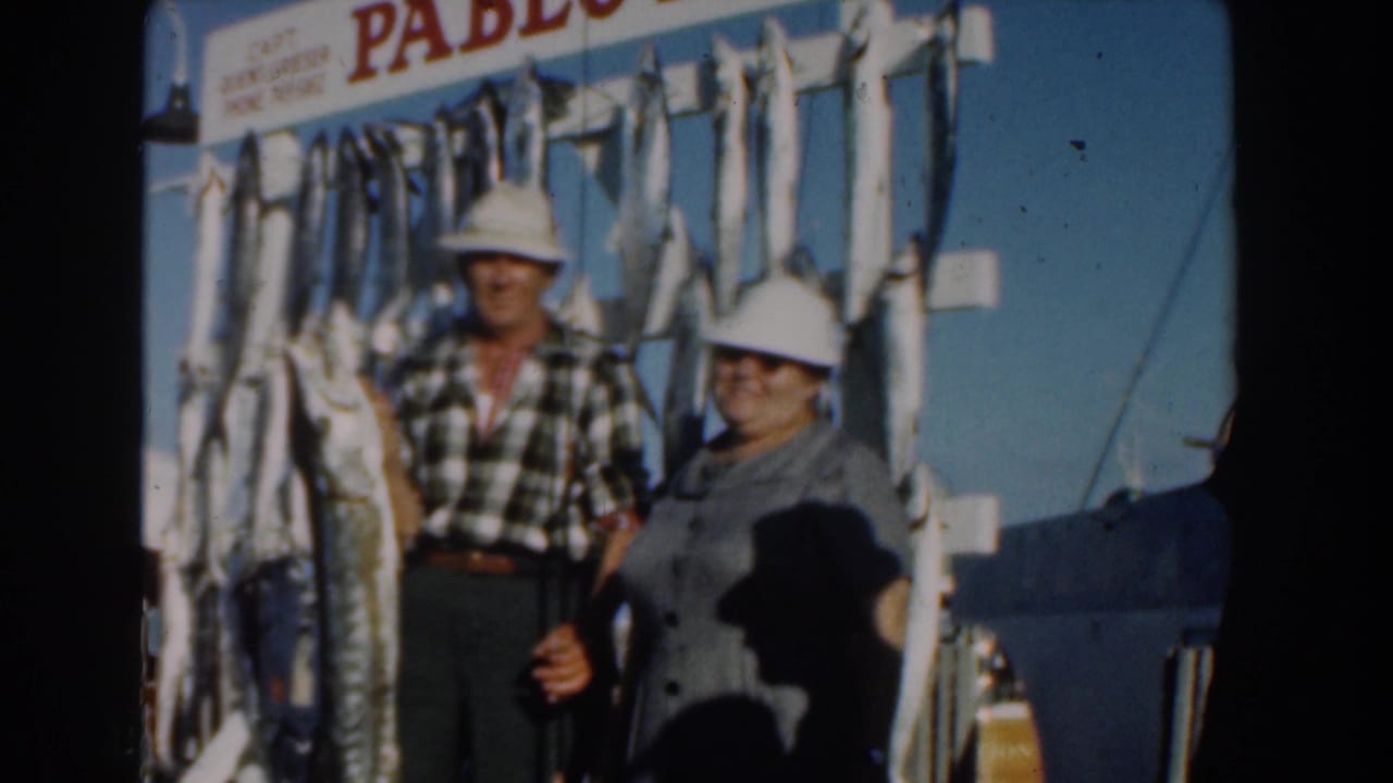 Elderly couple in sun hats pose beside large hanging fish on a boat.