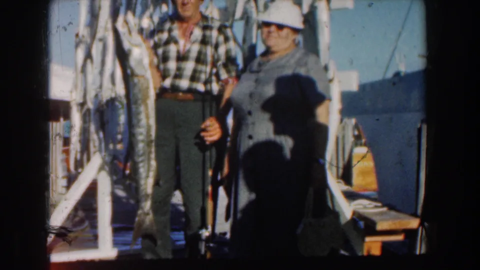 A closer 1961 Nassau home-movie frame shows the couple smiling beside a row of large fish hung on the boat.