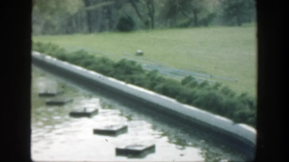 1949 Cincinnati home-movie frame of a landscaped pool with stepping stones, clipped shrubs, open lawn, and trees in the background.