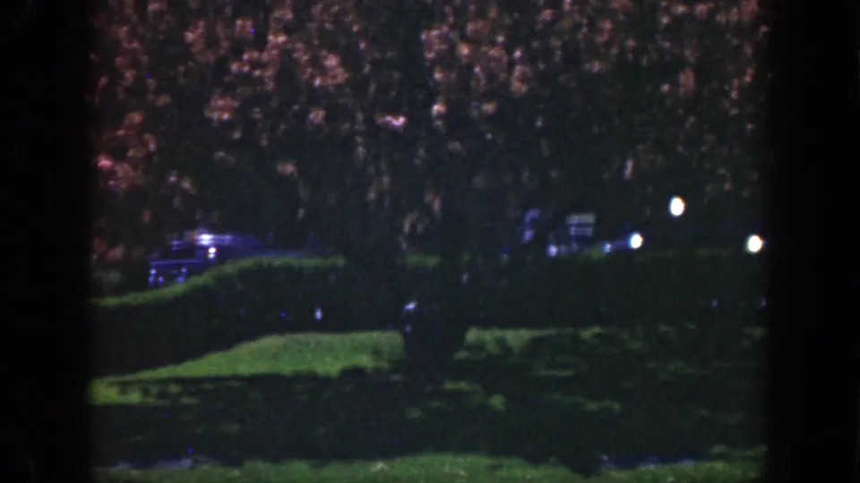 Evening-toned 8mm frame from the 1949 Cincinnati clip showing dark hedges, flowering branches, small lights, and a possible parked car.