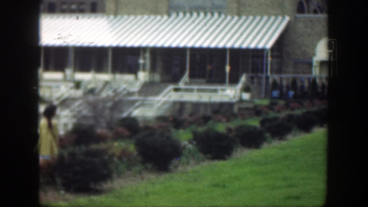 Tan brick building with striped awning, green lawn, shrubs, and a person in yellow at left.