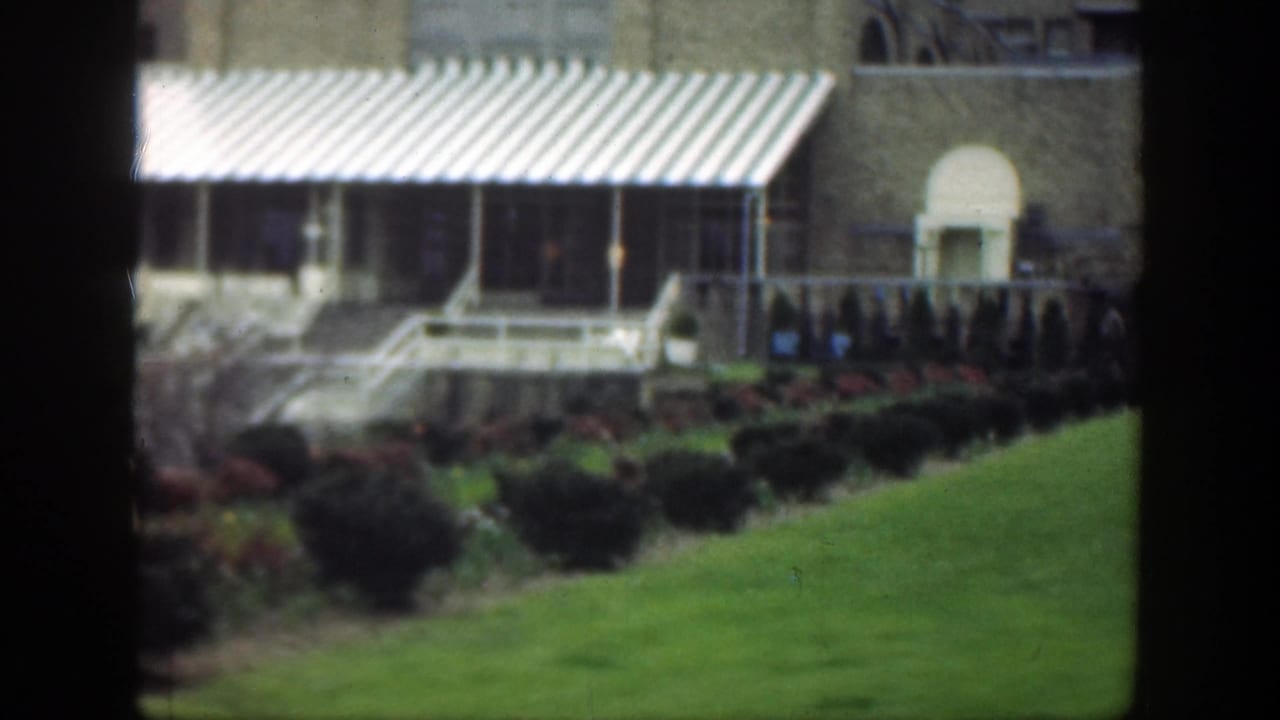 1949 Cincinnati exterior showing striped awnings, a tan brick facade, lawn, shrubs, steps, and a pale arched doorway.