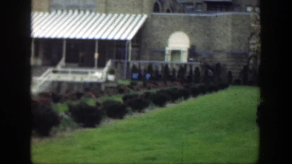 1949 Cincinnati home-movie frame with a sloping lawn, shrub border, striped awning, steps, and an arched doorway on a tan building.