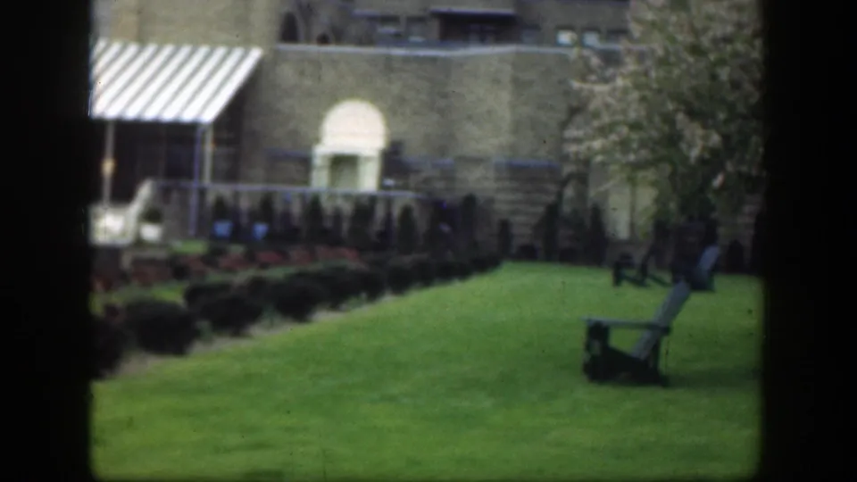 1949 Cincinnati exterior with a striped awning, lawn, shrub border, tan facade, arched doorway, and outdoor chairs at right.