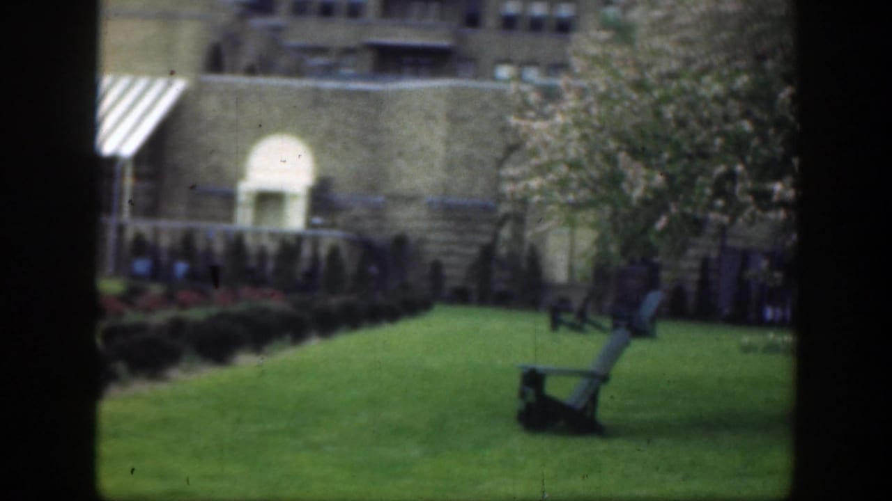 1949 Cincinnati home-movie frame showing a tan brick building, green courtyard lawn, flowering branches, and a wooden lawn chair.