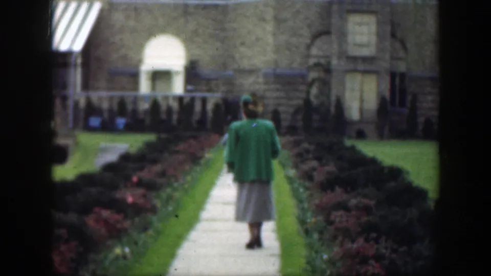 1949 Cincinnati frame showing a woman in a green jacket and light skirt walking along a courtyard path toward a tan building.