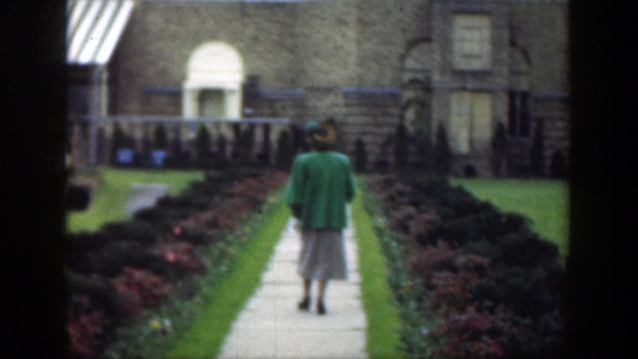 In 1949 Cincinnati home-movie footage, a woman in a green jacket and light skirt walks along a formal garden path toward a stone building.