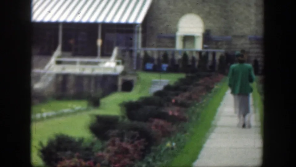 A home-movie frame shows the woman farther along the path, with hedges, lawn, railings, and a stone building in the background.