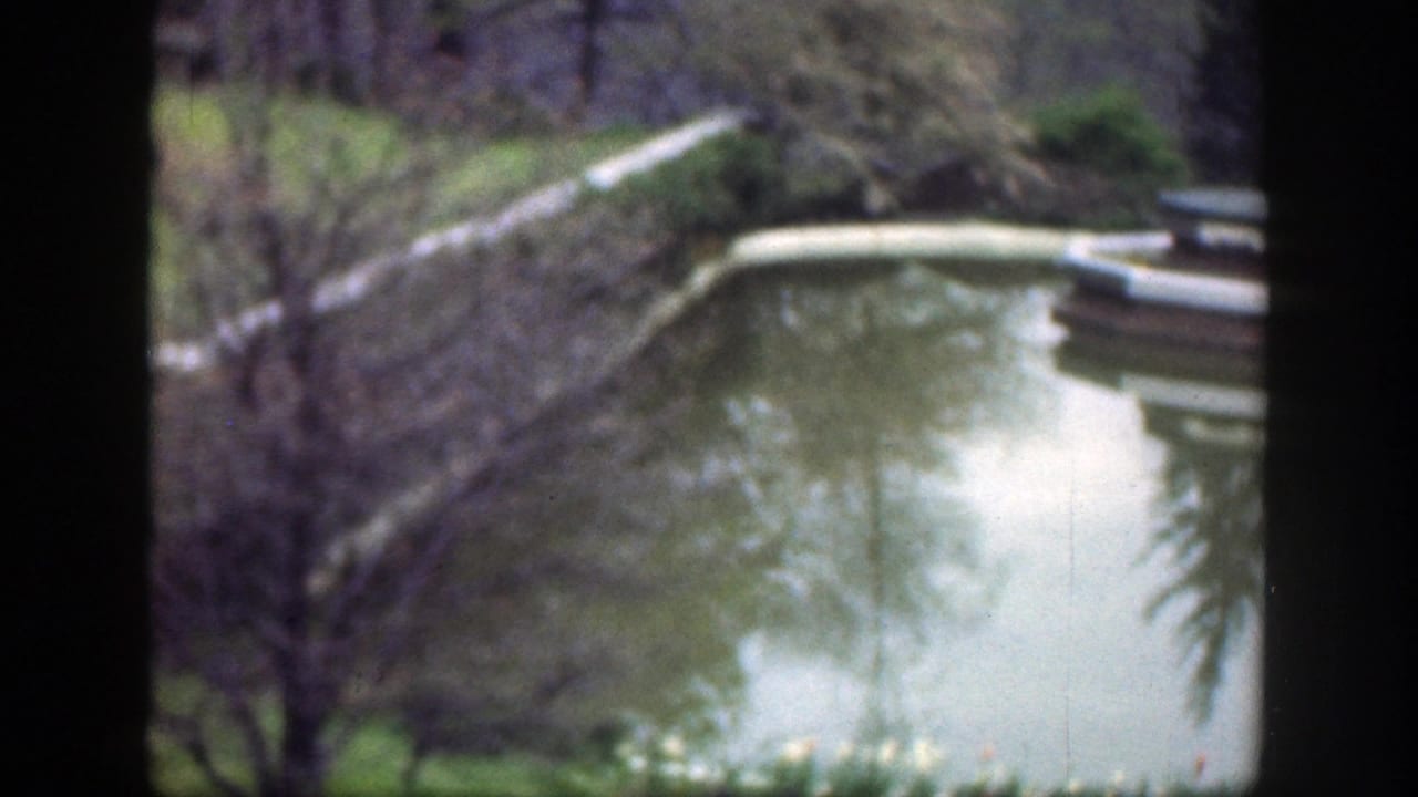 1949 Cincinnati-area home-movie frame showing a calm park pond, reflected trees, green grass, and a pale curved water feature.