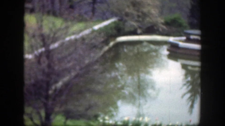 1949 Cincinnati-area home-movie frame showing a calm park pond, reflected trees, green grass, and a pale curved water feature.