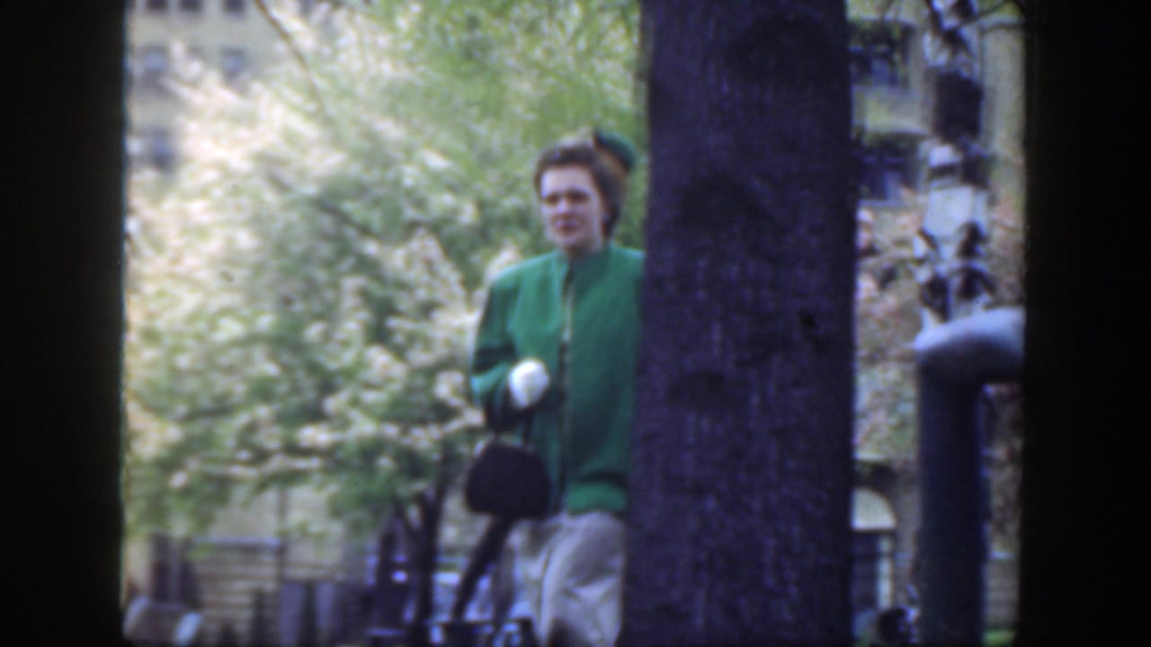 In 1949 Cincinnati, a woman in a green coat and hat walks past a tree with a large building behind her.