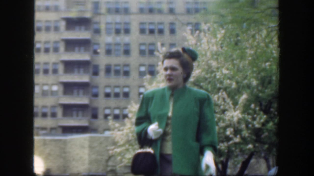 Woman in green coat holding a handbag with apartment building behind her.
