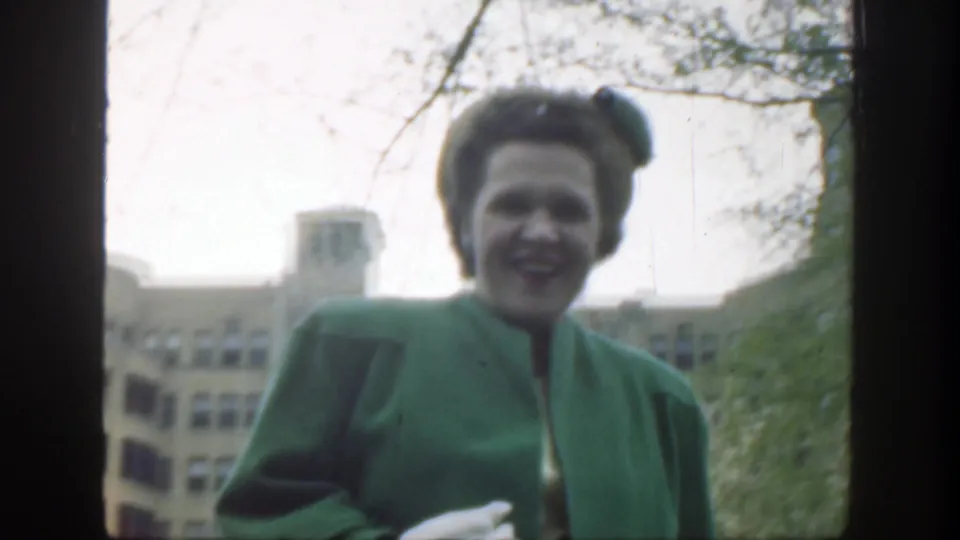 The woman smiles near the camera in a green coat and hat, with trees and a city building behind her in Cincinnati.