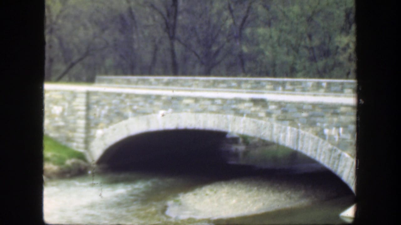 Stone arch bridge over a shallow river with trees behind it.