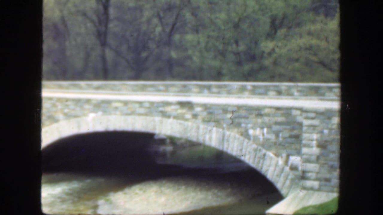 Stone bridge arch and railing seen across a tree-lined river.