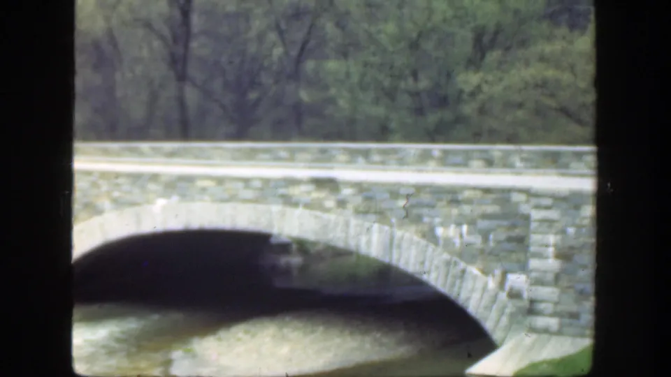 1949 Ohio home-movie frame with the bridge arch centered over the river and soft green trees beyond the road rail.
