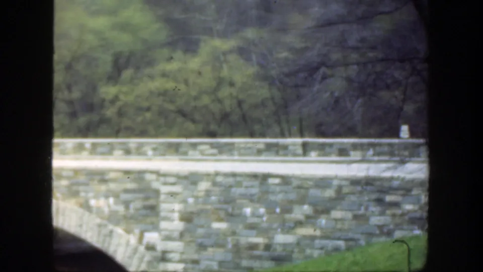 1949 Ohio home-movie frame showing the bridge wall and railing, wooded background, grassy bank, and partial arch at lower left.