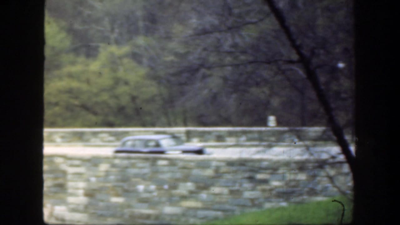 Dark foreground framing with a vintage car crossing a stone bridge beside trees.