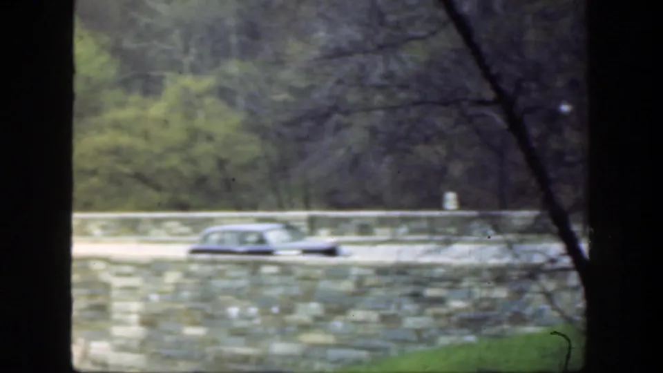 1949 Cincinnati home-movie frame showing a dark vintage car on a stone bridge, partly obscured by tree trunks and branches.