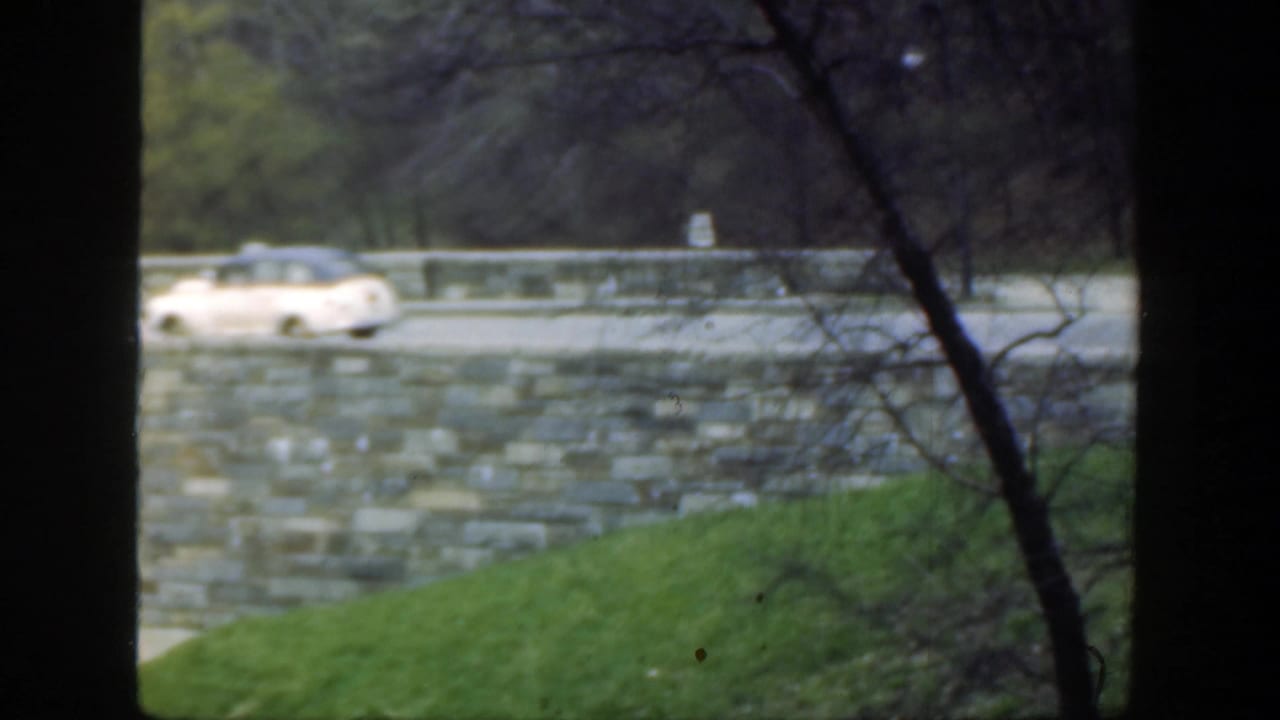 Light-colored vintage car crossing the bridge behind dark tree framing.