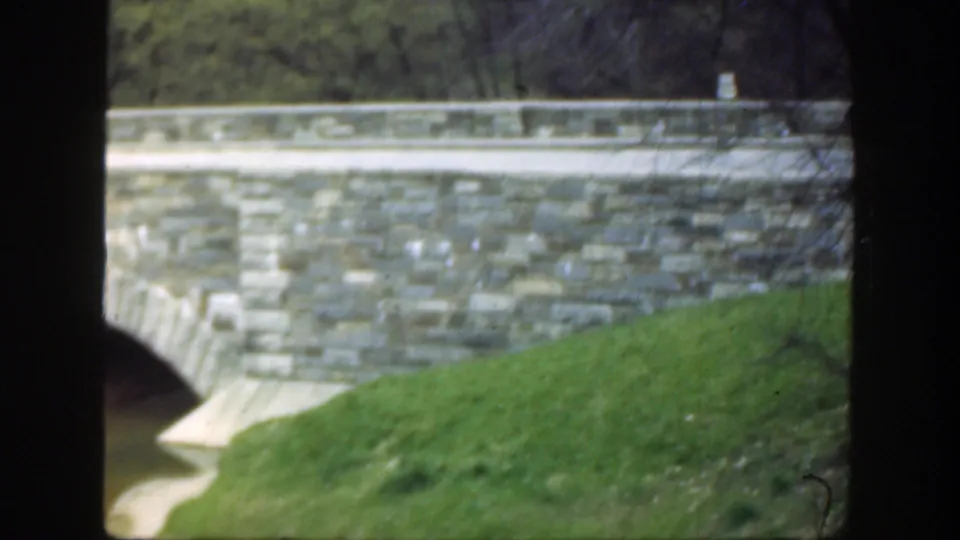 Stone arch bridge in Cincinnati home-movie footage, with water below, grassy slopes, and dark foreground framing.