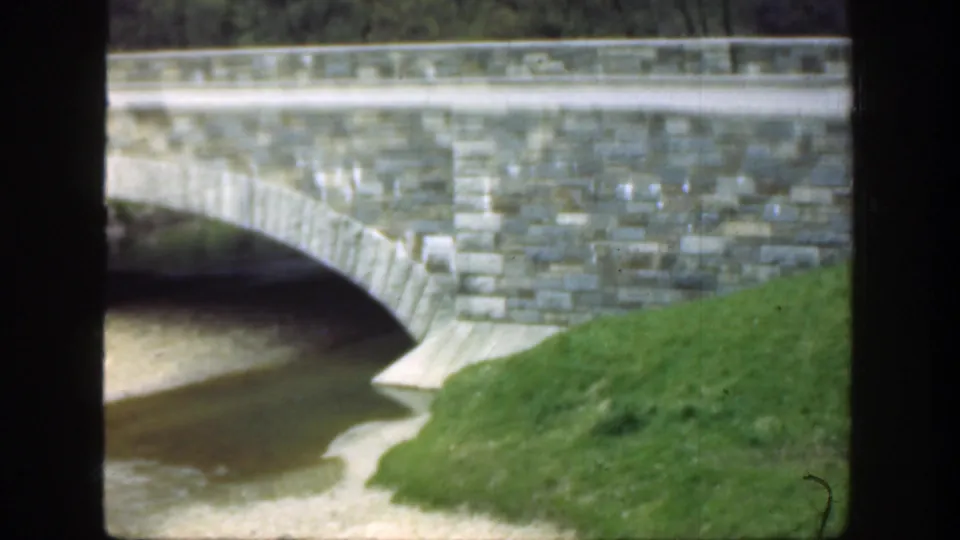 Late-1940s Cincinnati bridge view with a stone arch, calm water below, grass banks, and no visible people or signage.