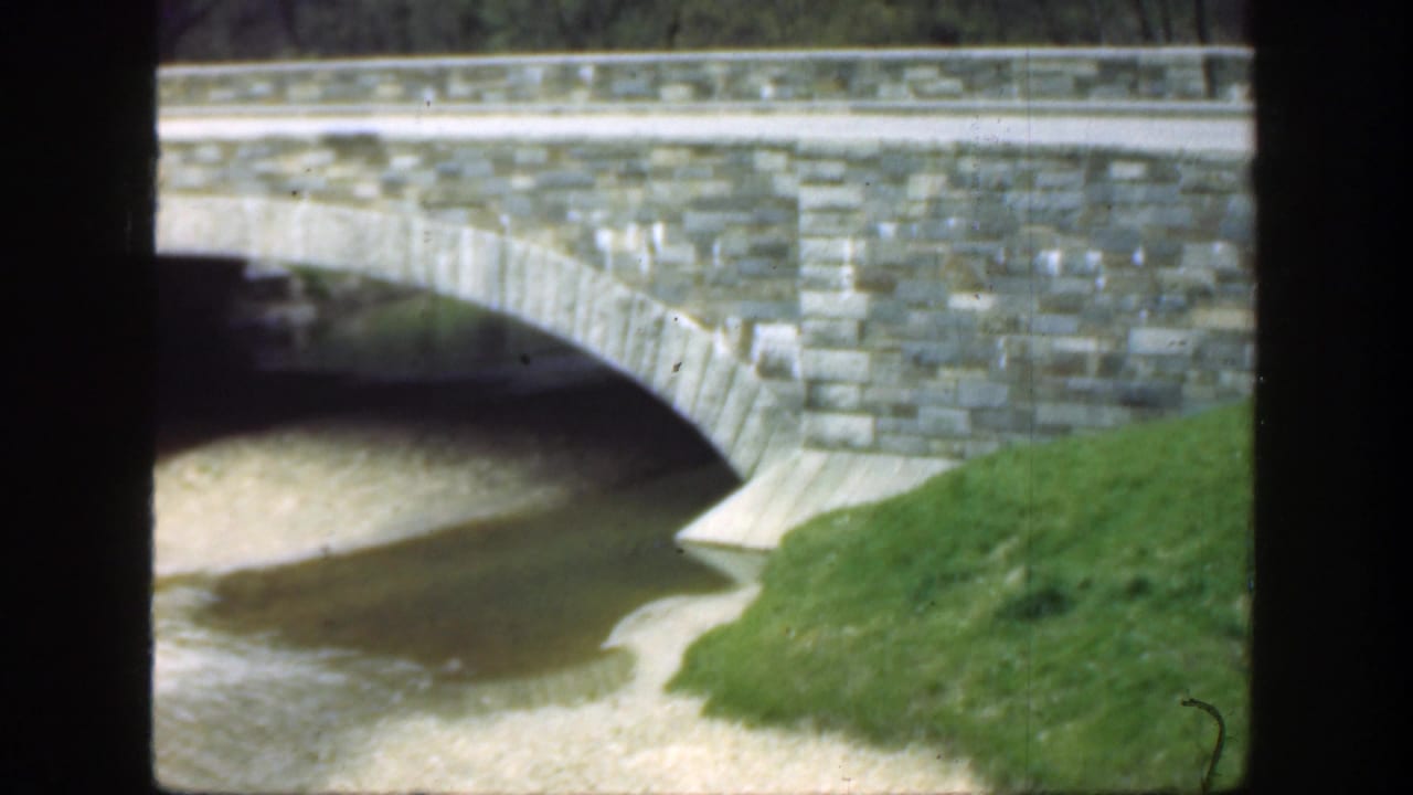 Gray stone arch bridge over shallow water with green grass in the foreground.