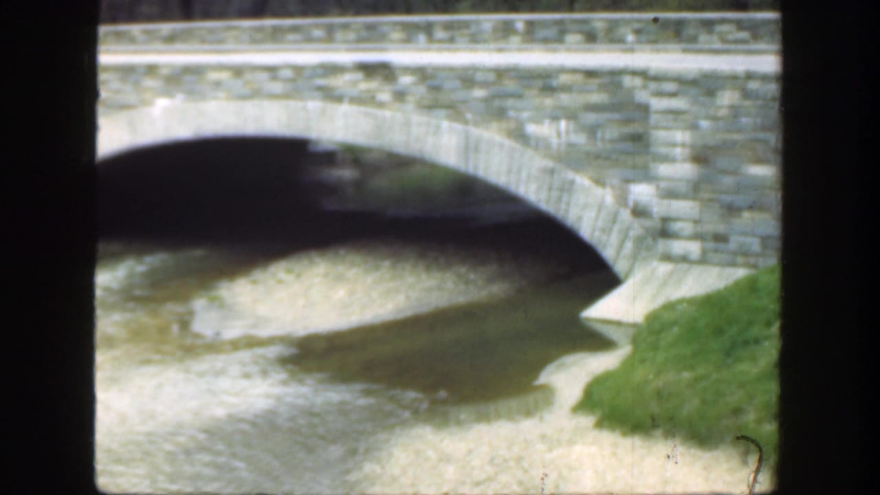 Stone bridge arch spanning shallow water with grass along the right bank.