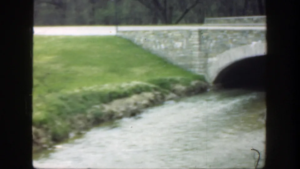 1949 Cincinnati frame with a grassy bank, shallow river, and part of the stone bridge at the edge of the view.