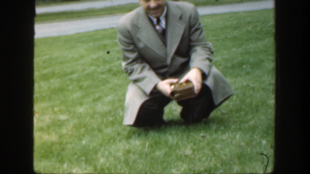 Adult man in a suit kneels on grass while holding a small box.