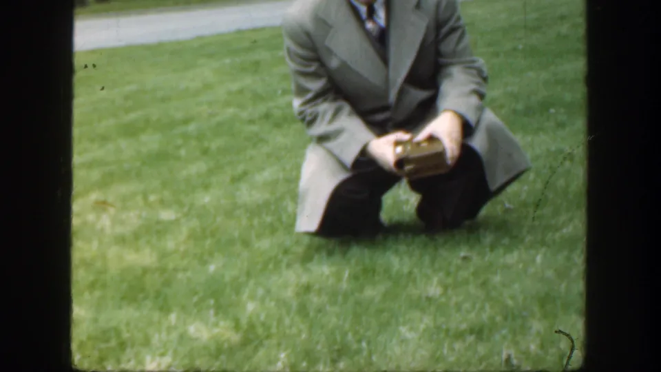 Cropped 1949 home-movie frame of a suited adult man kneeling on green grass and looking down toward the box in his hands.