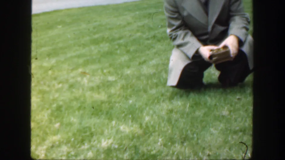 Final 1949 8mm frame shows the suited man partly cropped while kneeling on grass with the small box still in his hands.