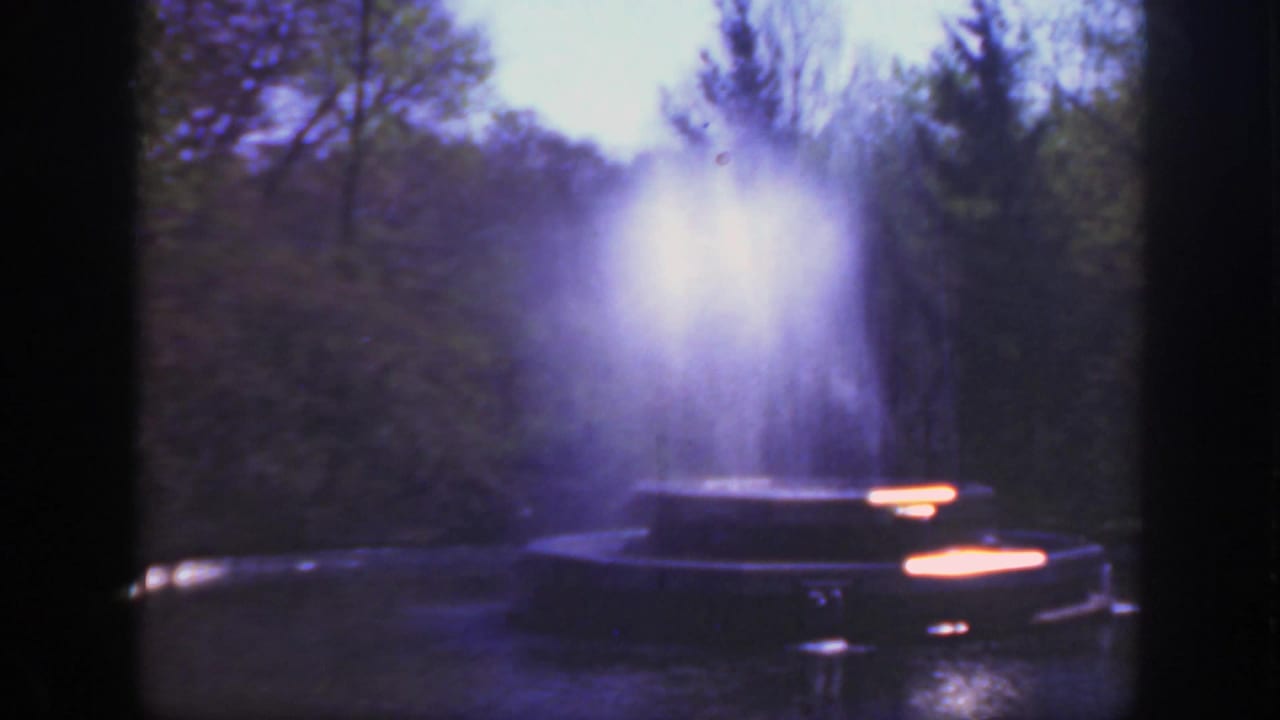 Park fountain with water spraying upward, trees behind it, and bright highlights on the basin.