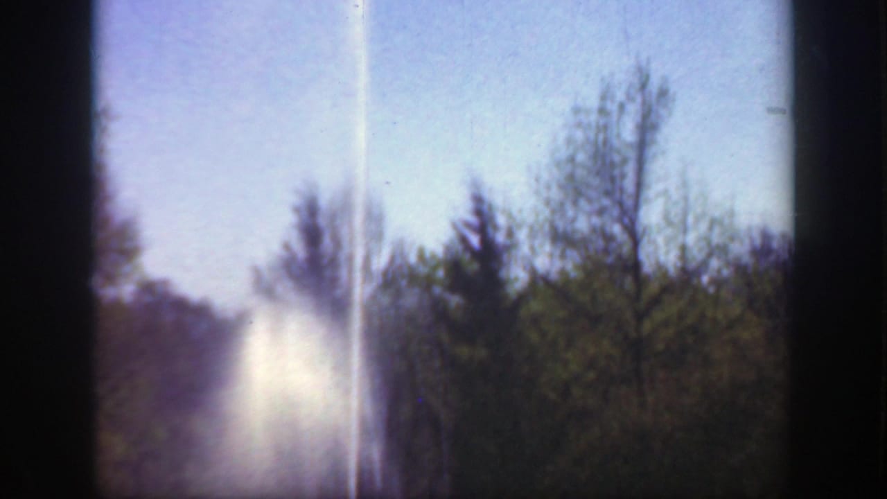Bright vertical beam or spray rises against a blue sky behind trees near the horizon.