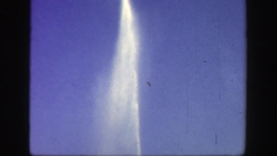 The 8mm frame centers on a bright vertical plume or light streak against blue sky, with film grain and edge shadows visible.