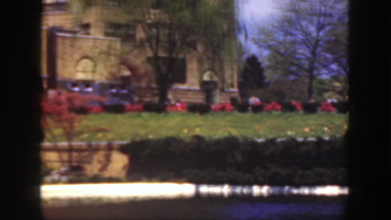 1949 Cincinnati 8mm still showing part of a stone mansion, manicured lawn, red flower beds, trees, and reflected water.