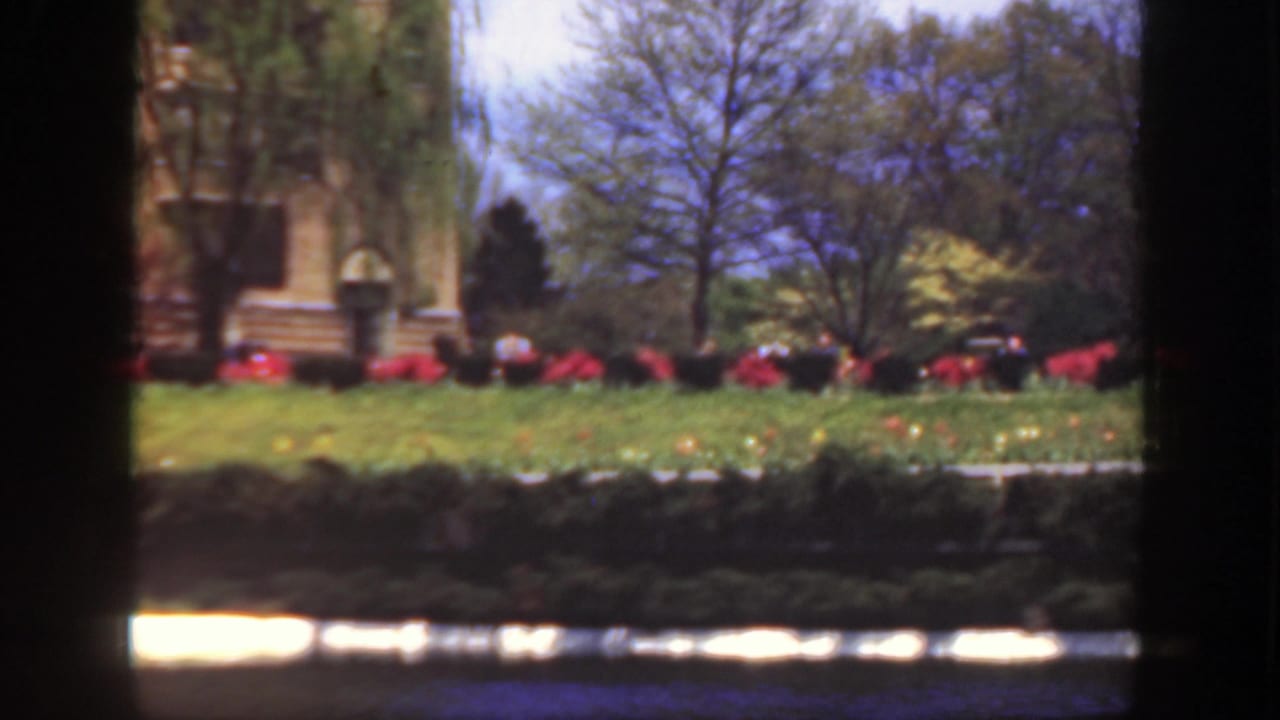 Formal lawn with red flower beds, trees, and part of a stone building behind dark vertical borders.