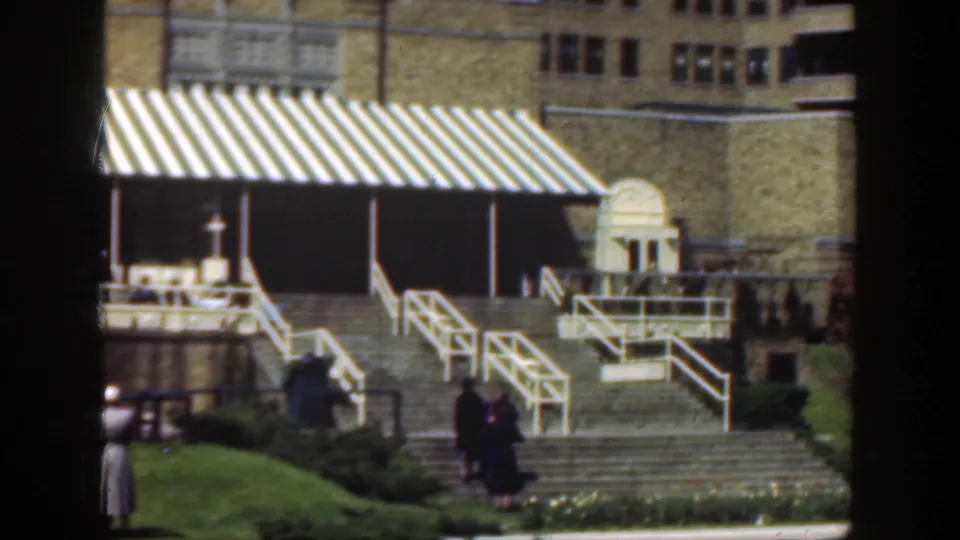 1949 Cincinnati exterior frame with white-railed steps, a striped awning, manicured lawn and two distant pedestrians in period dress.