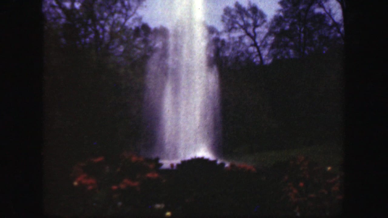 1949 Cincinnatti, Ohio 8mm frame of a tall park fountain rising above red flowers, with dark trees against a pale sky.