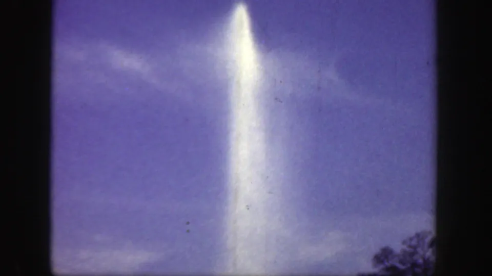 A bright column of water rises through the left-center of the frame, with purple-blue sky, wispy cloud, and dark treetop silhouettes visible in the 1949 Cincinnati home-movie footage.