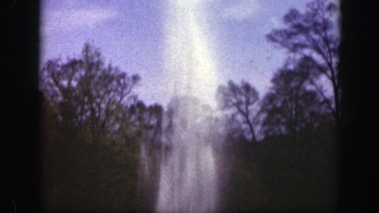1949 Cincinnati home-movie frame showing a tall fountain spray beyond a tree line and lawn, with soft sky and dark film edges.