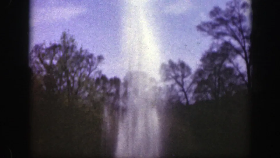 1949 Cincinnati home-movie frame showing a tall fountain spray beyond a tree line and lawn, with soft sky and dark film edges.