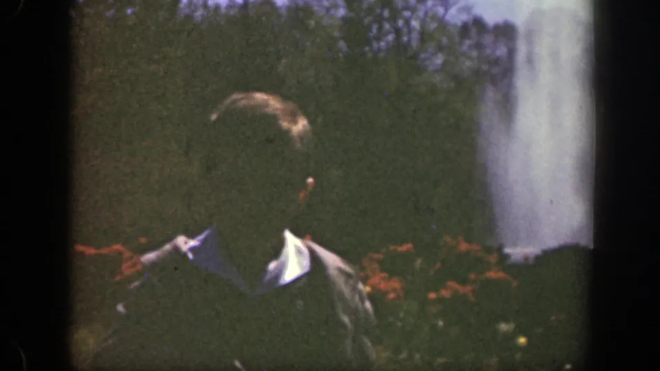The young boy is closer to camera in the Cincinnati park, framed with red flowers, green lawn, trees, and a large fountain plume.