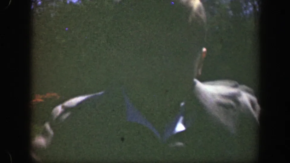 A close, dark 8mm frame shows the boy in a suit jacket and light shirt, with the park fountain and tree line behind him.