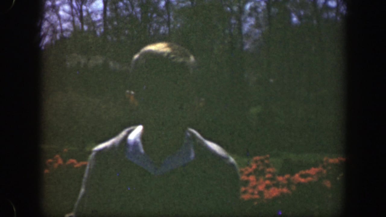 In 1949 Cincinnati home-movie footage, a young man in a tan jacket stands in a sunlit garden with red flowers behind him.