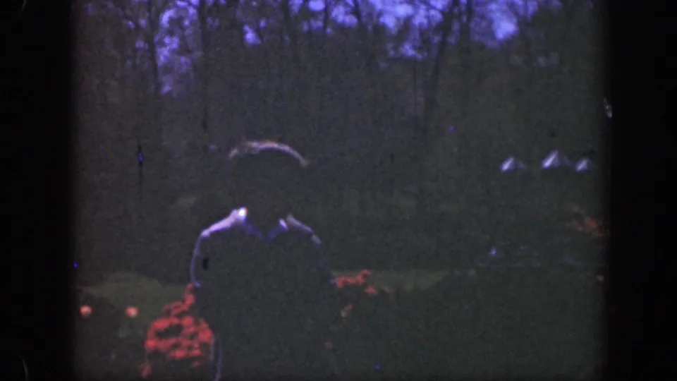 A shadowed man in a tan jacket faces the camera in a Cincinnati garden, with red flowers and dark trees behind him.