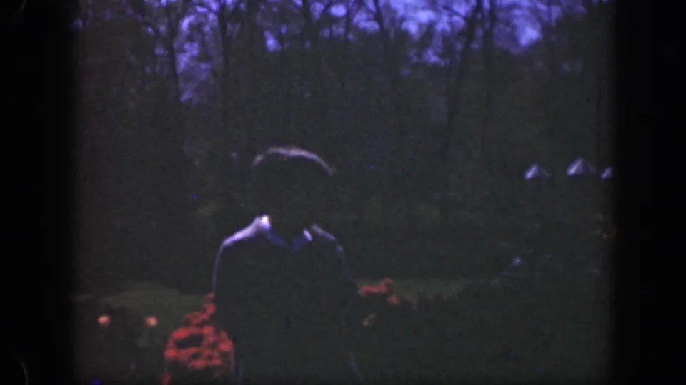 A dark, backlit figure approaches through a Cincinnati garden, with red flowers and trees visible in soft 8mm focus.