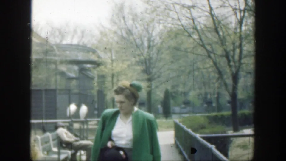A woman in a green jacket walks through the Cincinnati zoo area, with benches, trees, fencing, and birds or pale objects in the background.